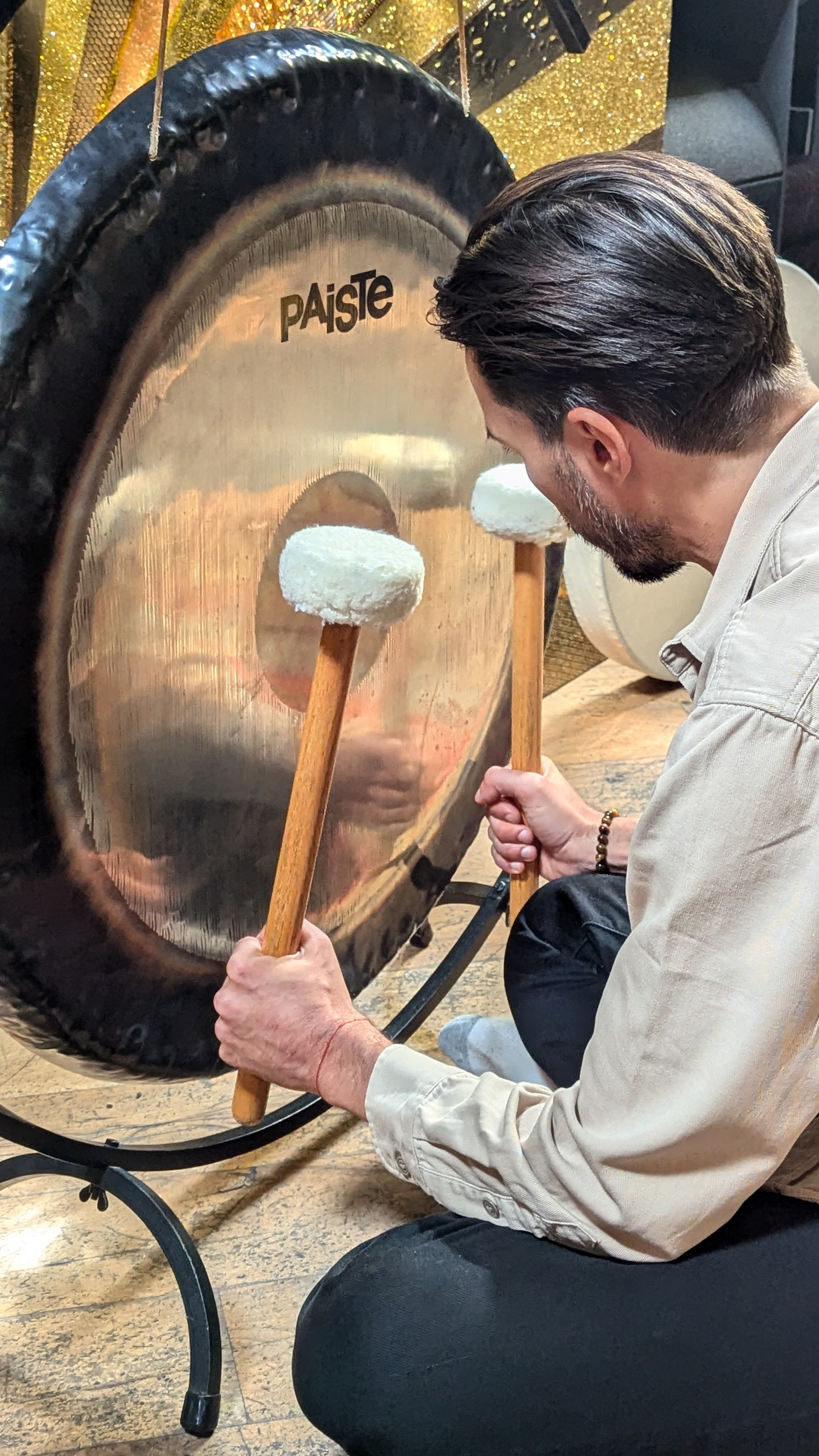 Savino seated cross-legged before a bronze Paiste gong, felt mallets in hand, warm golden bokeh light behind