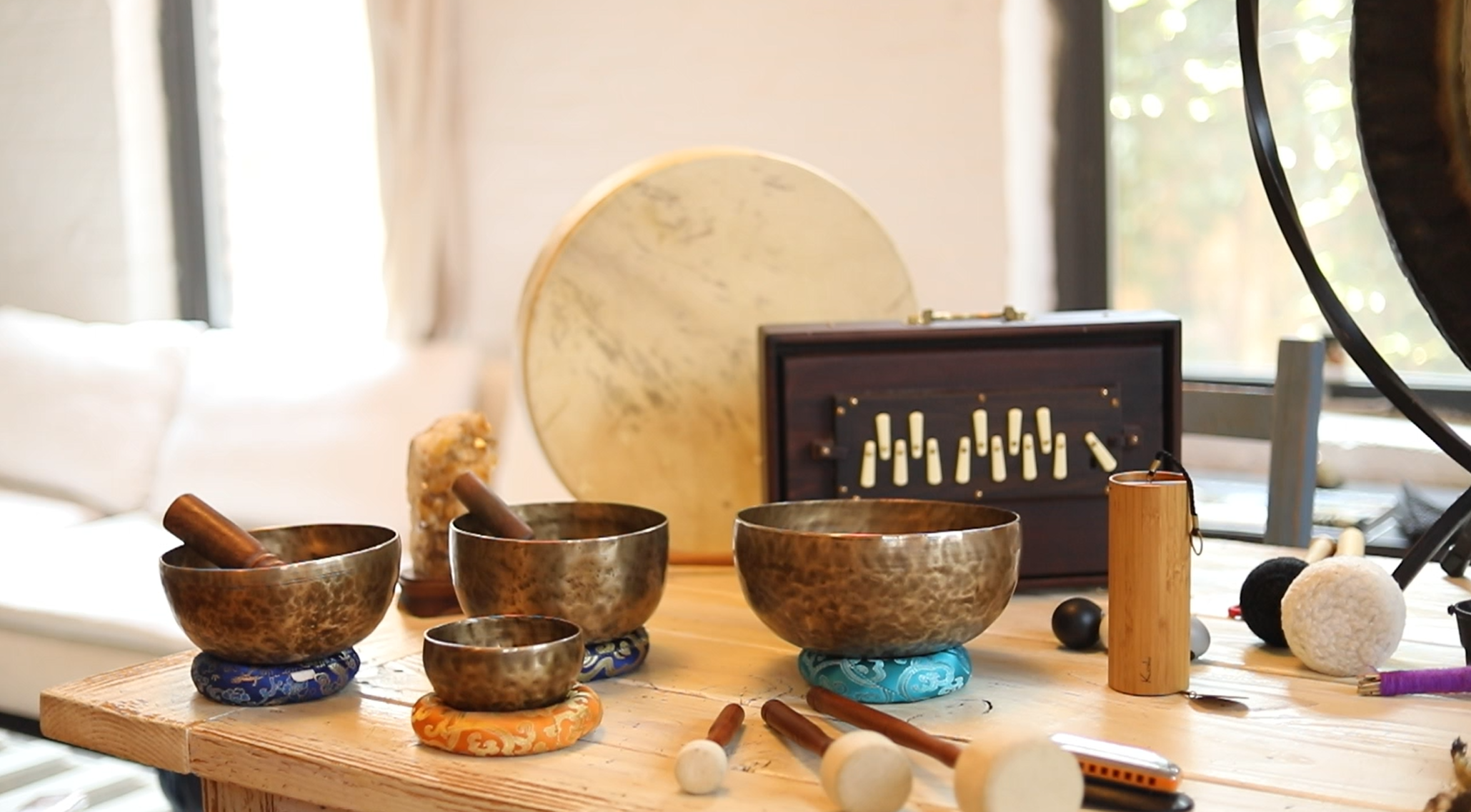 Collection of Tibetan singing bowls, shruti box, frame drum, and chimes arranged on a wooden table in natural light